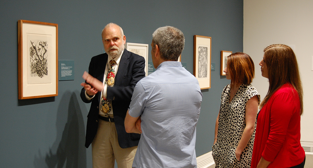 Curator talks to museum visitors in front of drawings hanging on a blue wall