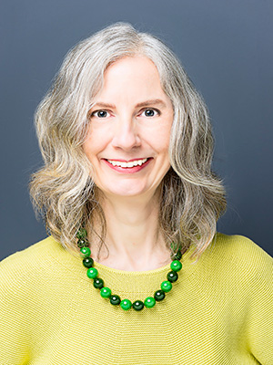 A woman with light hair and skin wearing a yellow sweater and green necklace
