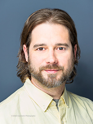 A headshot of a man with brown collar-length hair, a moustache and beard, white skin, and a light green collared shirt