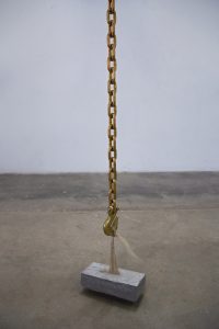 A long gold chain with a large hook holds up a cement block by an attached bunch of blonde hair