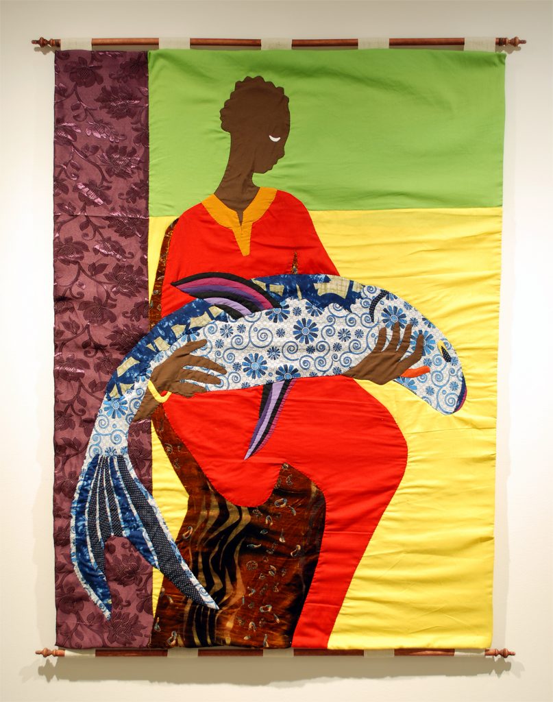 Tapestry with an androgynous Black person sitting in the center, holding a limp blue fish in their arms. The person’s face is in profile, and they are looking down towards the fish. The background consists of rectangular sections of fabric in colors of purple, green, and yellow. The person wears a robe of red, orange, and brown fabric. The fish consists mainly of a blue and white patterned fabric of flowers and swirls with pink, purple, and black fins.