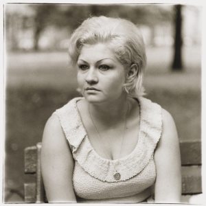 Black and white photo of a woman with platinum blonde hair and light skin wearing a scoop neck top and sitting on a park bench