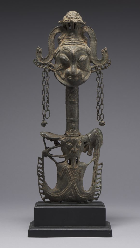 A metal rattle. The bottom section depicts a human with legs transforming into a mudfish, creating a U shape. Directly above that is a bird with a large beaked head on one side and its tail feathers on the other. Above the bottom section is a cylindrical handle. Above the handle is a double headed-figure. The figure displays slightly different but similar features and has an enigmatic smile. The figure wears a headdress at the top of which there appears to be hair. On each of the figure’s ears are two chains with bells attached. The rattles stands on a square base.