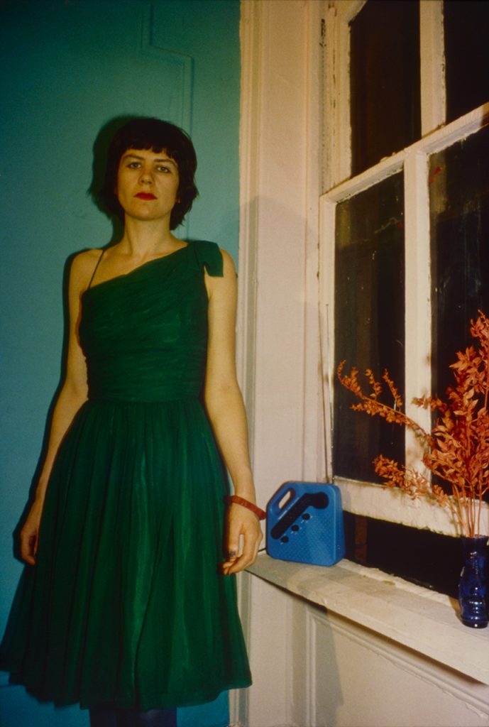 Color photo of a woman with dark hair and light skin wearing a green one-shoulder dress and standing by a window against a turquoise wall