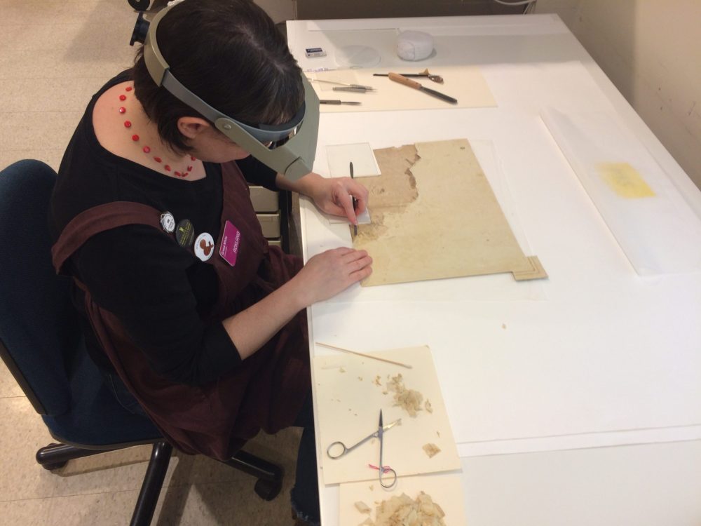 A paper conservator removes backing from a drawing using tweezers and scissors