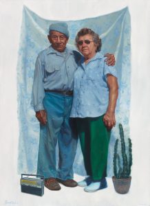 Two elders with short gray hair and light brown skin stand in front of a curtain beside a cactus and behind a transistor radio.
