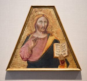 A light-skinned person with gold halo, beard, and draped tunic holding a book and making a sign of blessing with the right hand