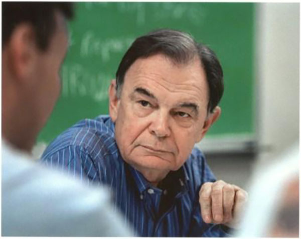 A man with light skin and short dark hair wearing a blue and white striped shirt sits in front of a green chalkboard