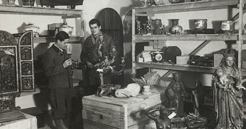 Two men in uniform with light skin and dark hair stand in a storage room filled with art.